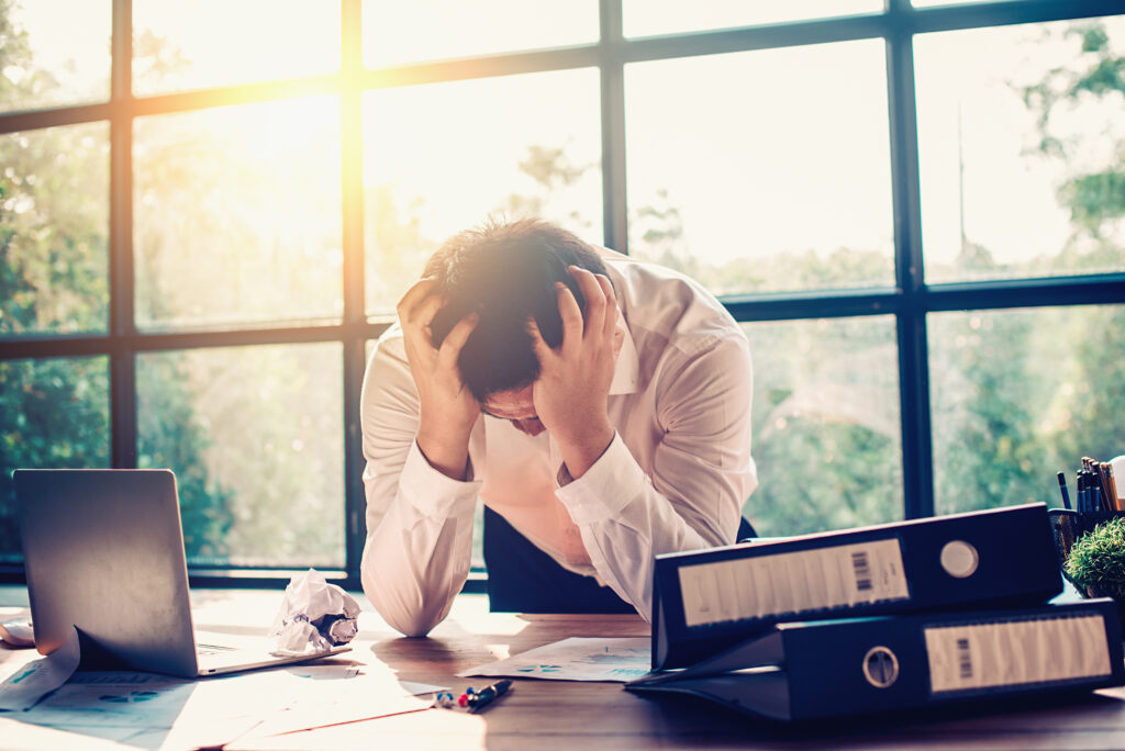 Overwhelmed man sitting at a desk with his head in his hands, surrounded by scattered papers and sunlight from large windows behind him.