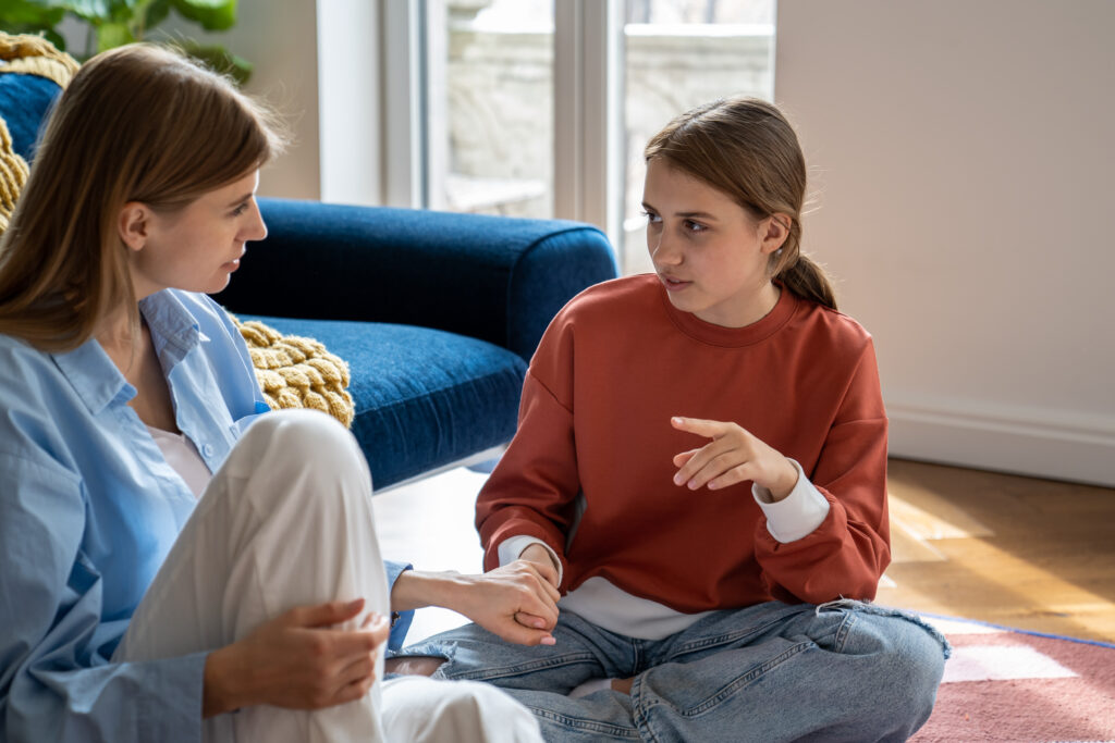 Two people sit on the floor facing each other and talk, a girl in an orange sweatshirt and a woman in a light blue shirt visible beside a blue couch.