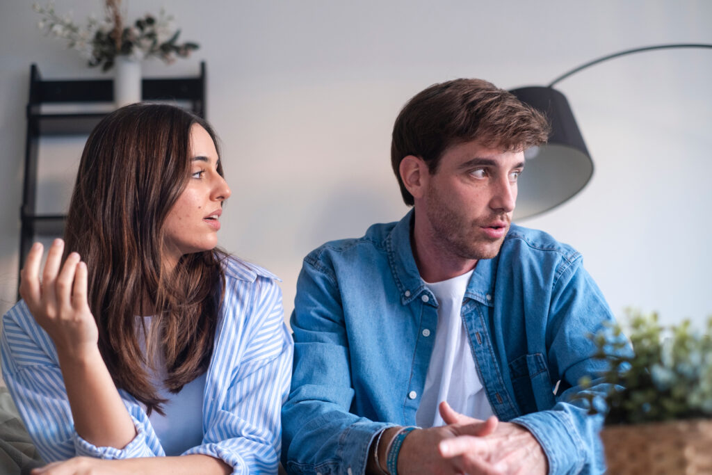 Two young adults sit at a table and talk; woman on the left gestures with her hand, man on the right listens.