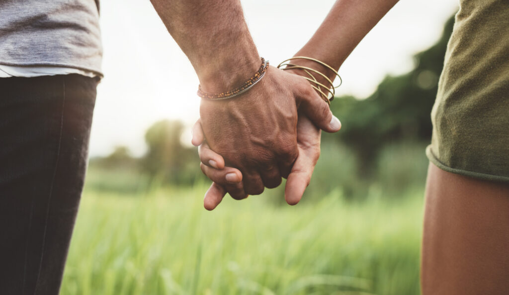 Two people hold hands in a sunlit field, showing affection and connection with bracelets on their wrists.